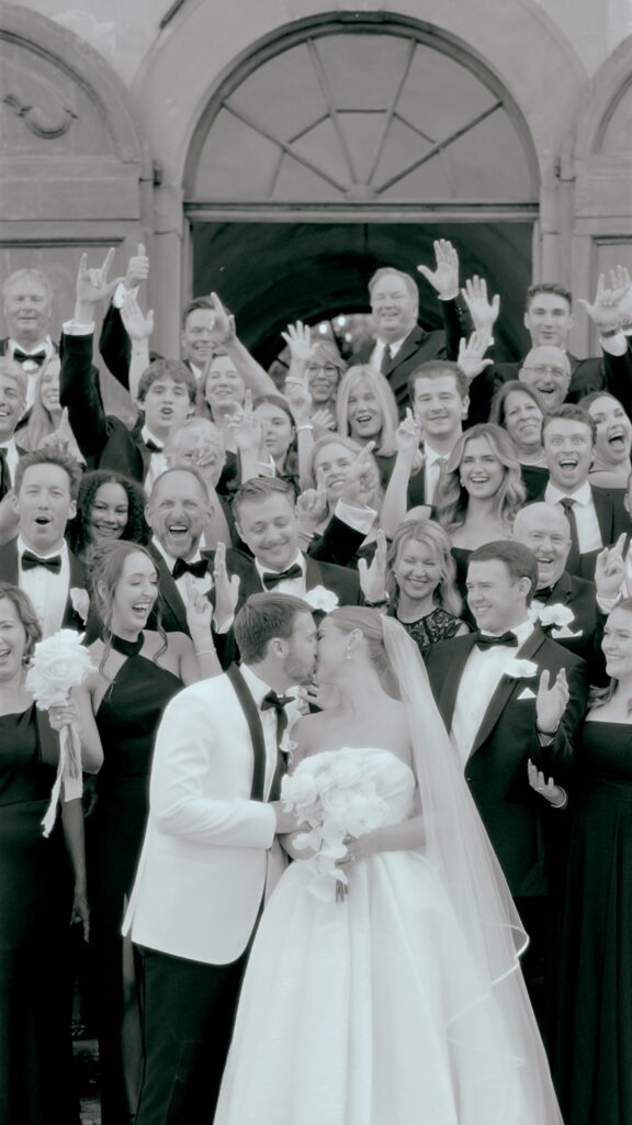 professional wedding videographer capturing the Bride and groom kissing in front of cheering wedding guests outside a historic building
