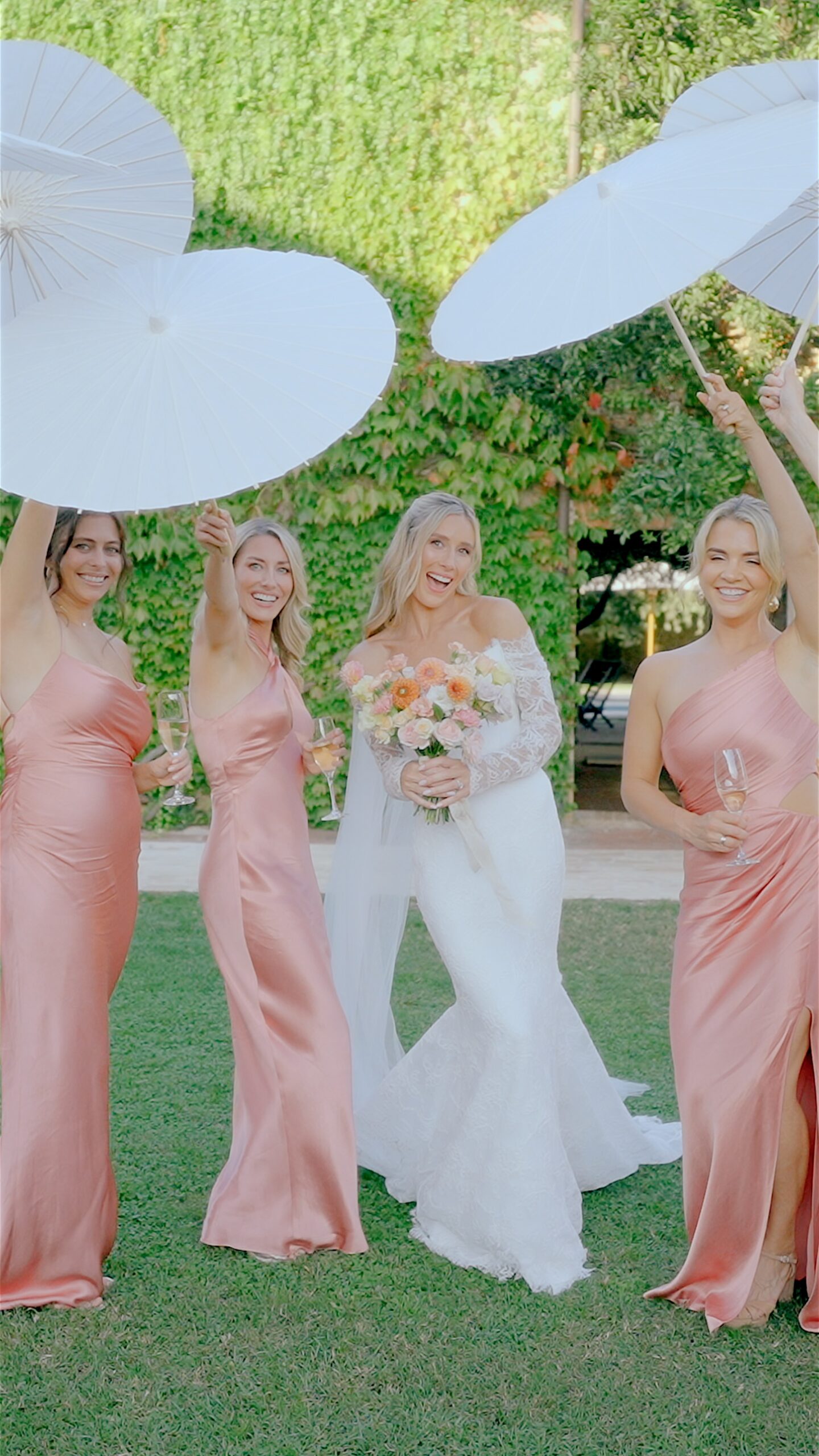Bride and bridesmaids in pink dresses holding white parasols and champagne glasses in a garden
