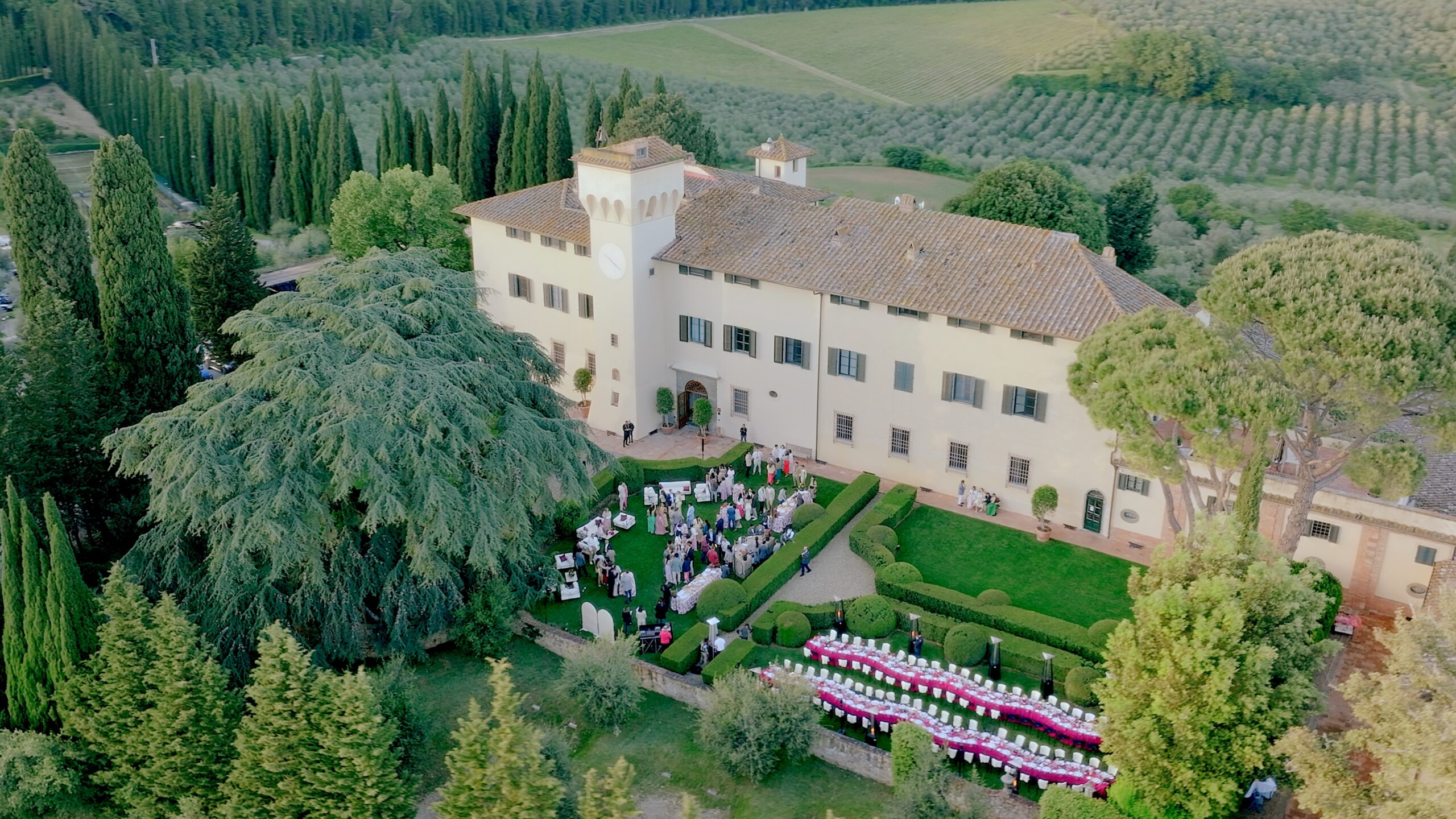 Aerial view of a historic Tuscan villa with wedding guests, long reception tables, gardens, and countryside surroundings