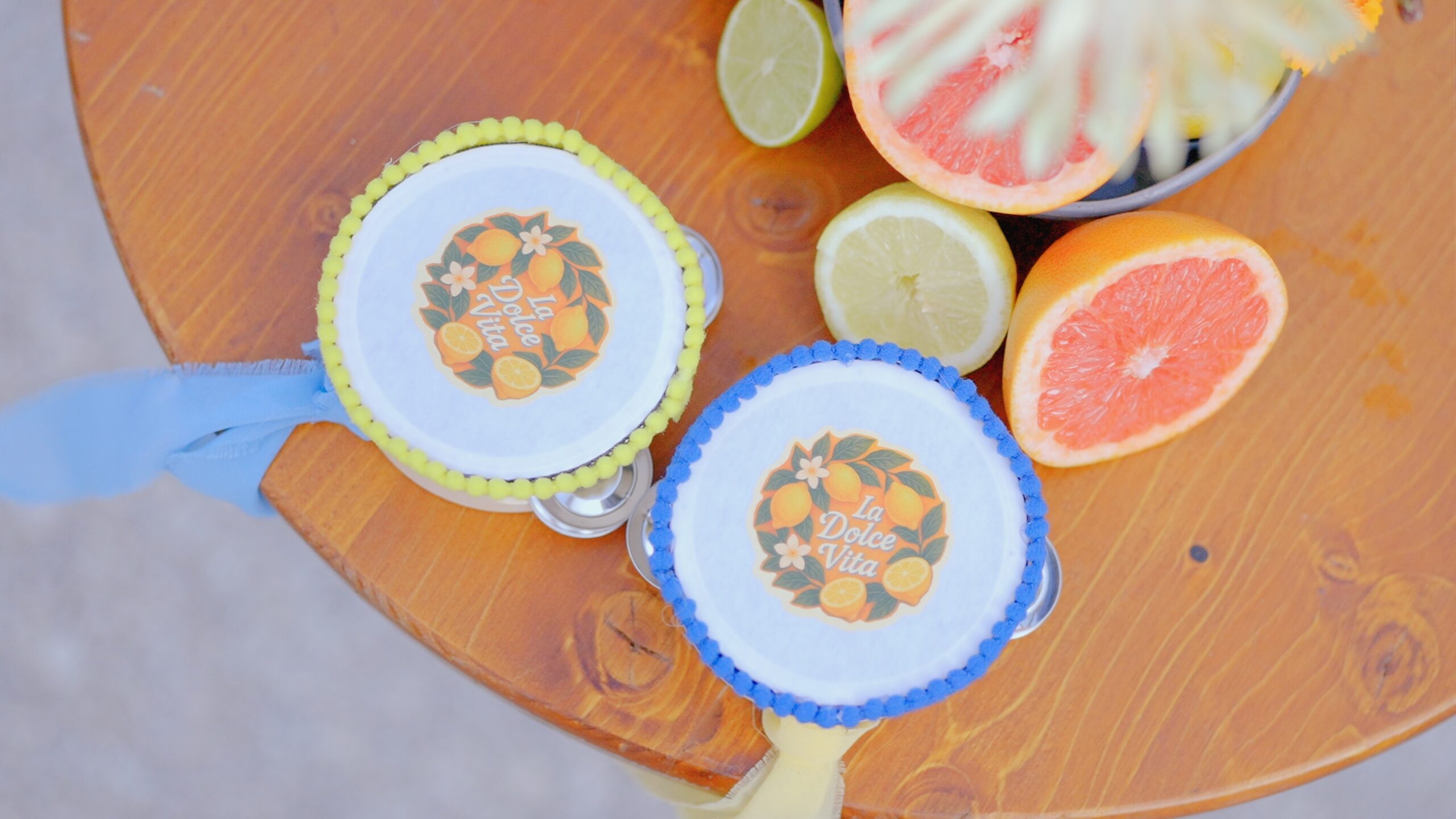 Two decorative tambourines with “La Dolce Vita” labels placed next to grapefruit, lemon, and lime slices on a wooden table