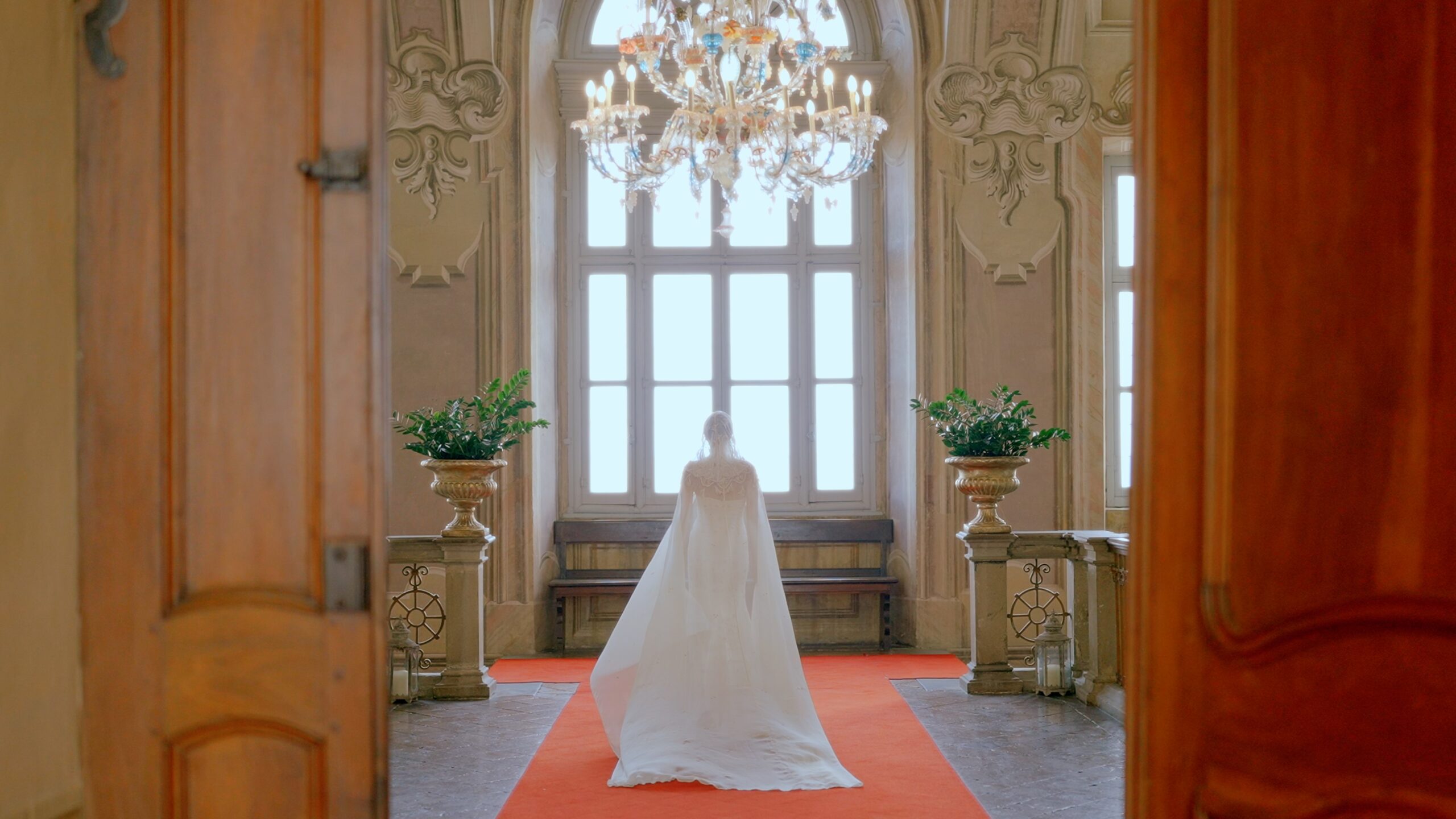 Bride standing alone in a grand historic hall with tall windows and a chandelier.