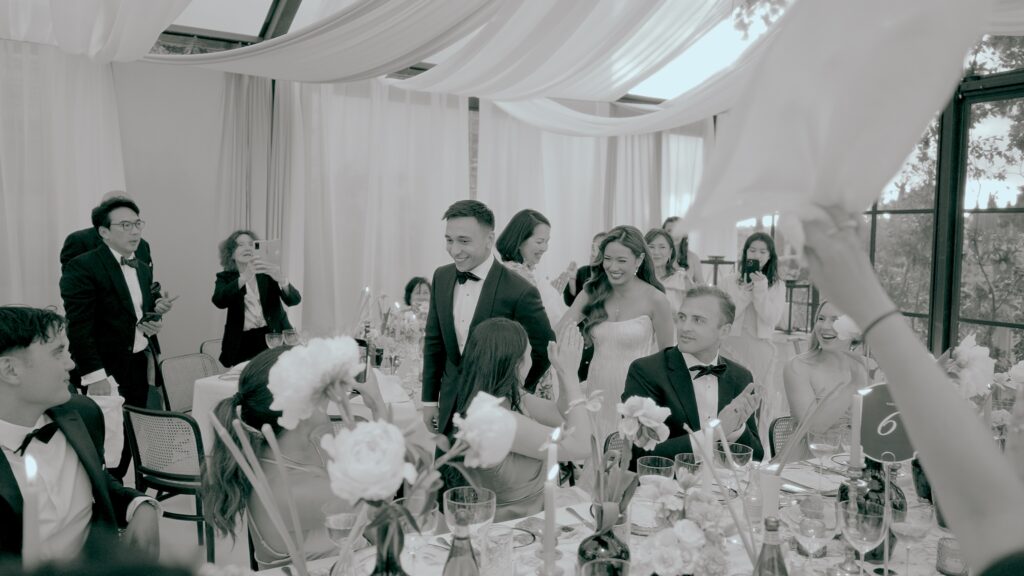 Bride and groom entering a stylish wedding reception while guests clap, smile, and take photos around decorated tables.