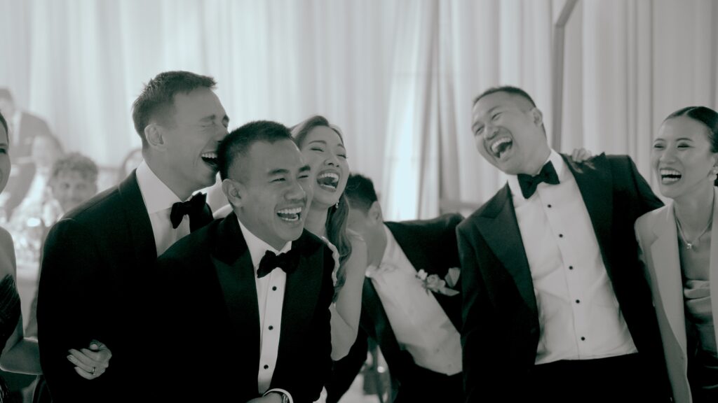 Wedding guests in tuxedos and formal attire laughing together during a stylish indoor reception.