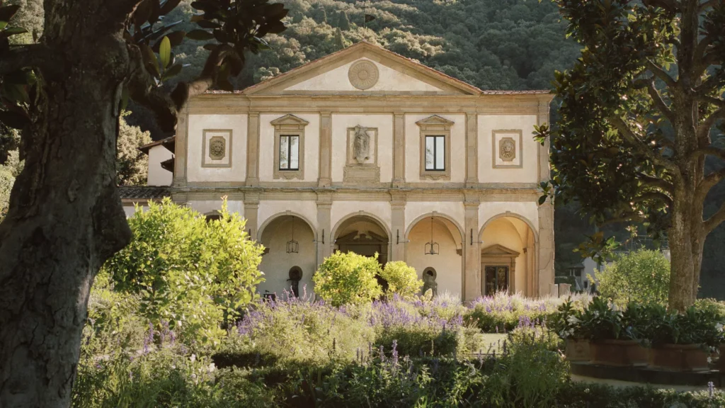 Historic Tuscan villa with arches, symmetrical façade, flowering gardens, lavender plants, trees, and wooded hills in the background.