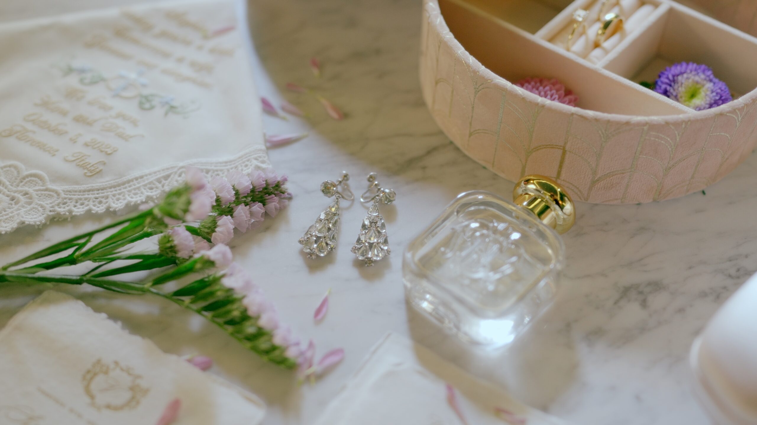 Bridal accessories including earrings, perfume, flowers, and a jewelry box arranged on a marble surface.