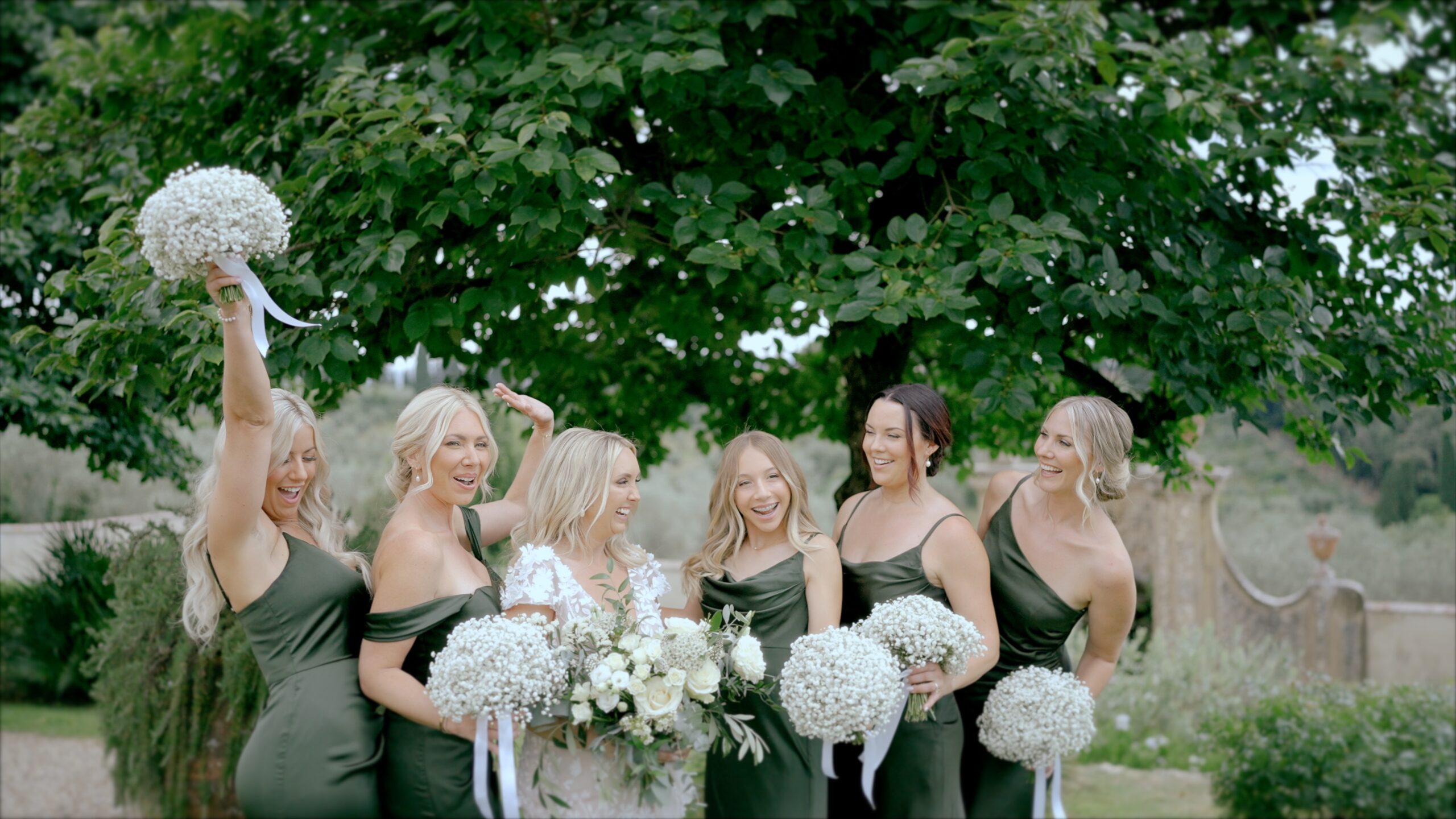 Bride and bridesmaids in dark green dresses laughing together outdoors with white flower bouquets.