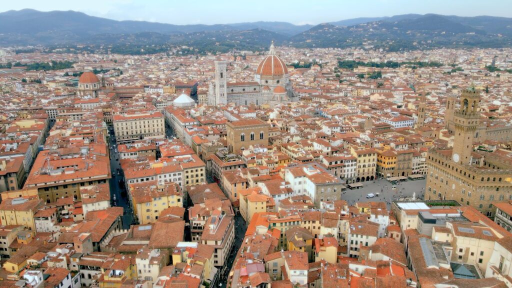 Aerial view of Florence with the Duomo, Palazzo Vecchio, terracotta rooftops, historic streets, and hills in the background.