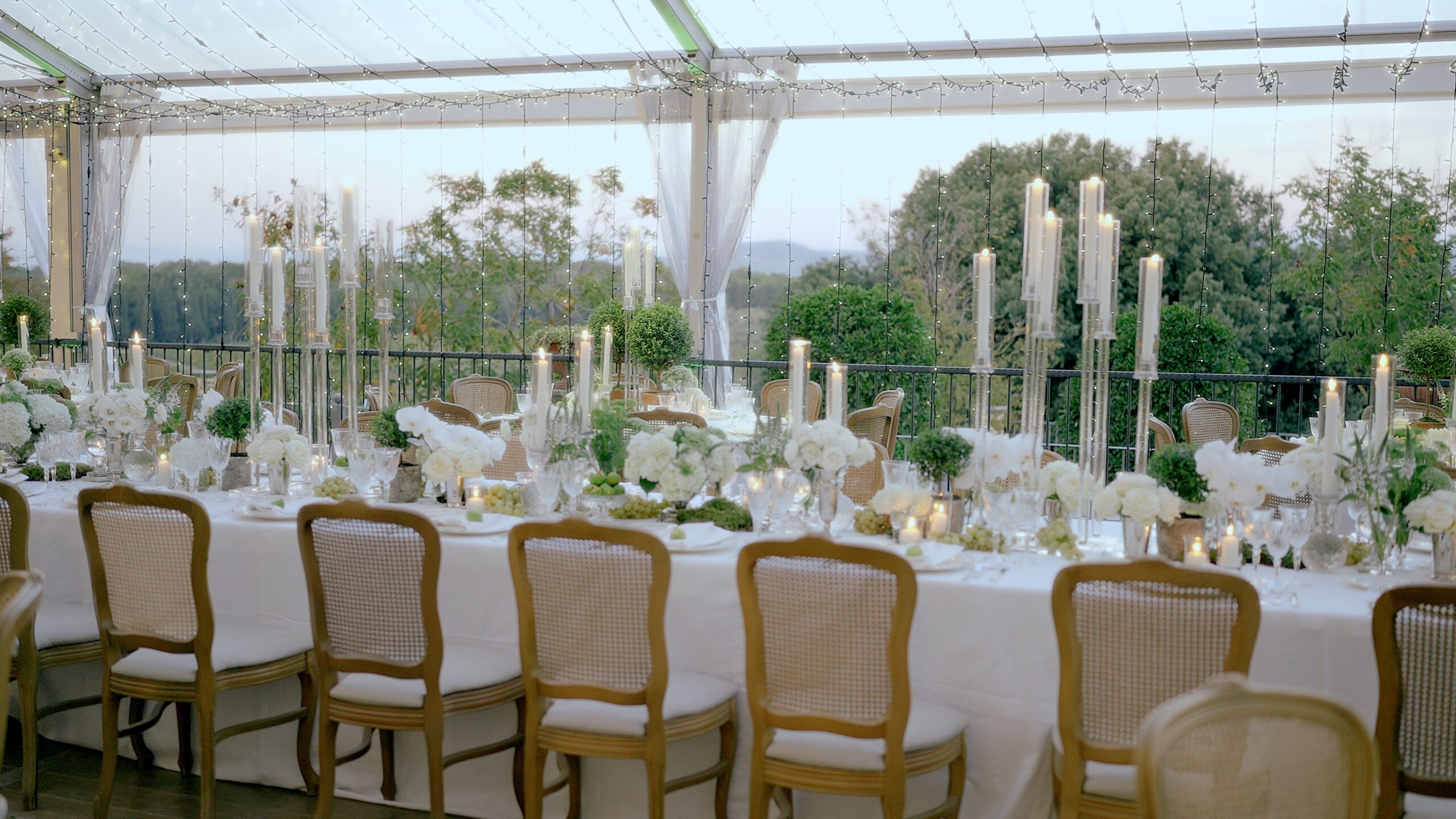 Elegant wedding reception setup with long tables, white flowers, tall candles, string lights, and cane-back chairs inside a glass marquee with countryside views.