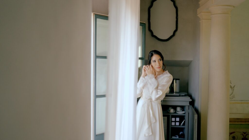 Bride in an ivory silk robe adjusting her earring in an elegant room with soft light, classic columns, and a mirror in the background.