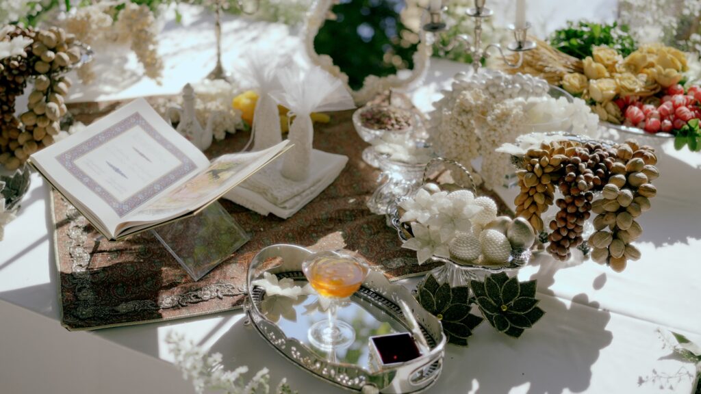 A sunlit wedding table decorated with white flowers, silver trays, candles, an open ceremonial book, sugared almonds, nuts, and a small drink in an elegant glass.