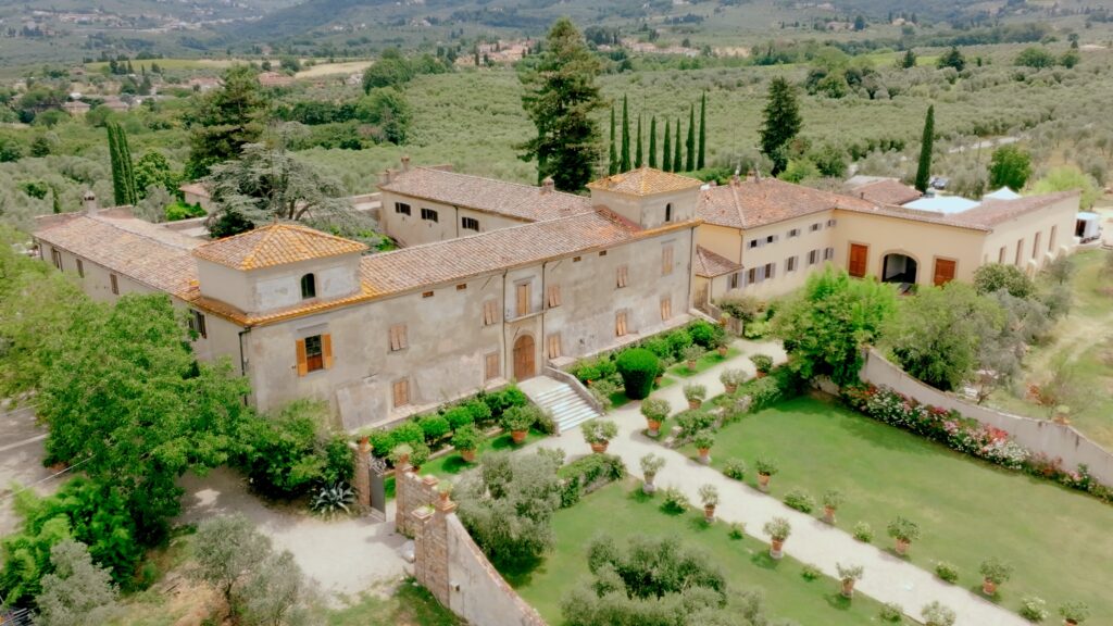 Large Tuscan countryside estate with stone buildings, terracotta roofs, green gardens, gravel paths, olive trees, and rolling hills in the background.