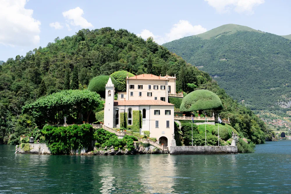 Exterior view of Villa del Balbianello on Lake Como, Italy, featuring historic architecture, manicured terraced gardens with umbrella-shaped trees, and the surrounding mountains and water.