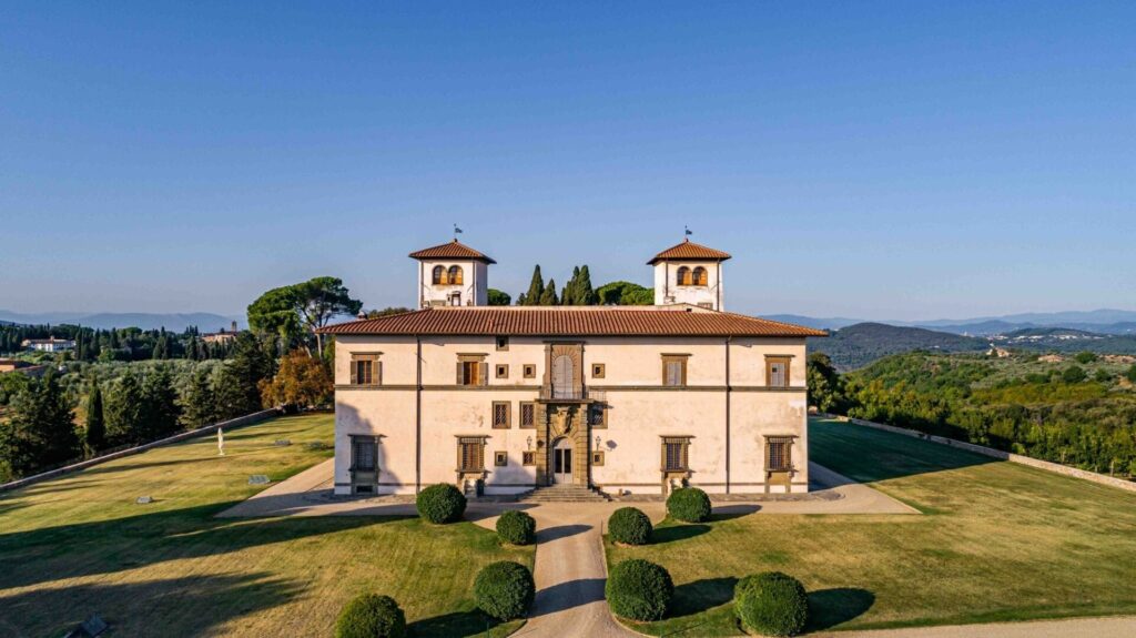 Large historic Tuscan villa with symmetrical architecture, terracotta roofs, round trimmed bushes, green lawns, and panoramic views of rolling hills and trees in the background.