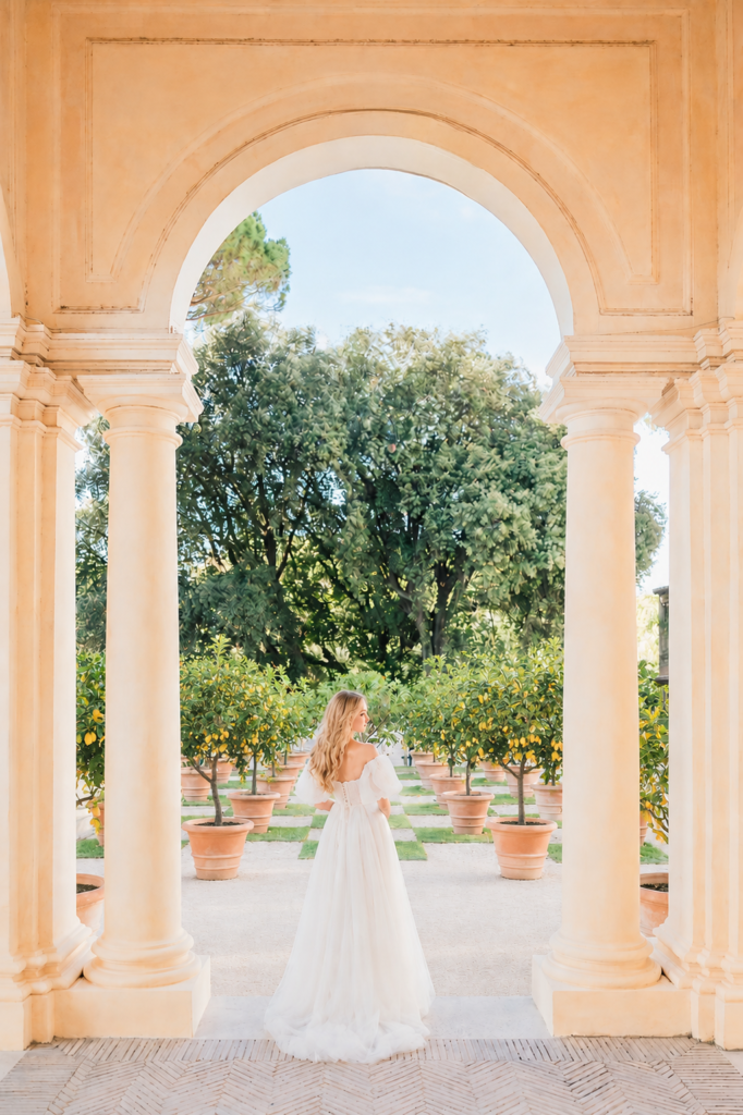 Amalfi Coast Wedding Videographer 3 Blonde bride in a flowing white wedding dress standing beneath an elegant Italian archway surrounded by potted lemon trees at a luxury Amalfi Coast wedding venue.
