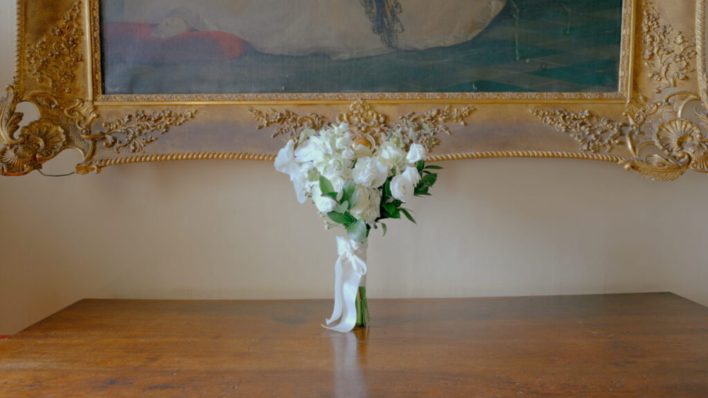 Why U.S. Couples Choose Italian Wedding Videographers 8 A luxury white bridal bouquet with roses and orchids tied with a silk ribbon, displayed on an antique wooden table beneath a large ornate gold-framed painting inside an Italian villa.