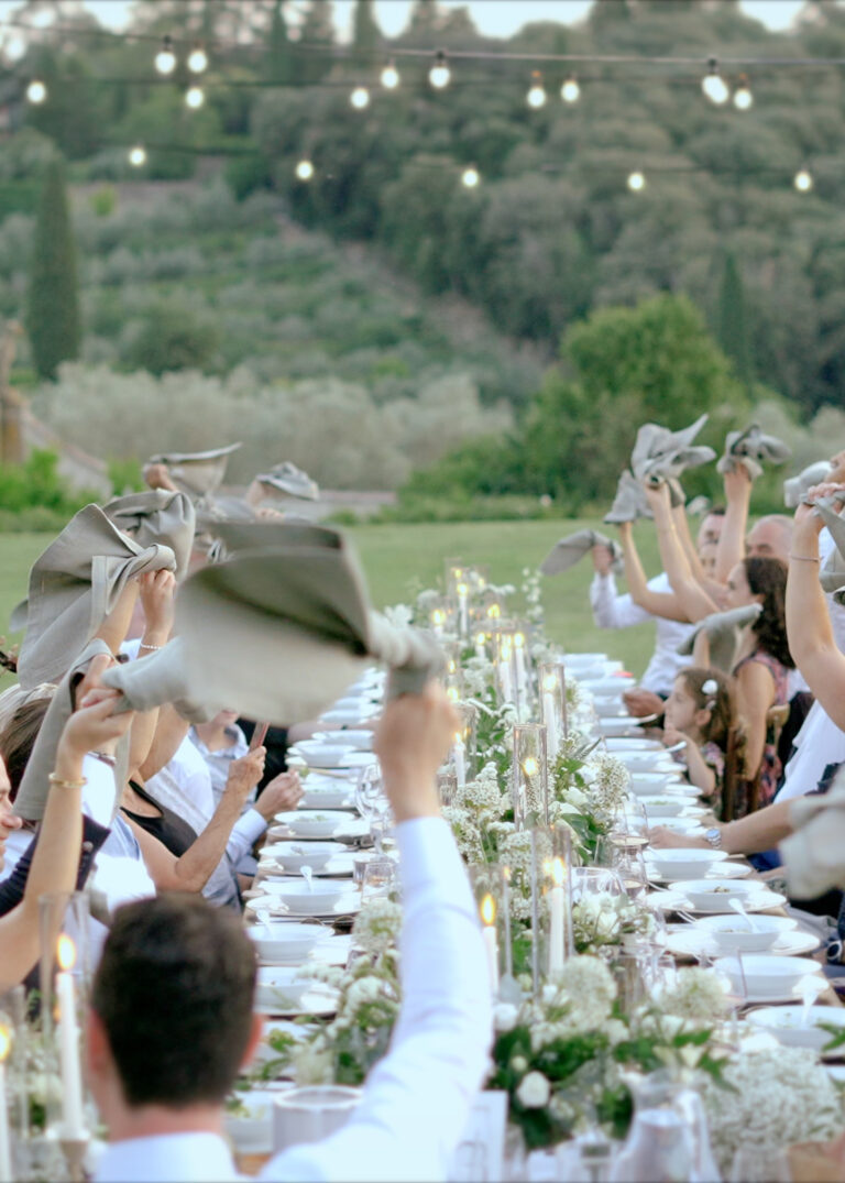 Destination Wedding Videographer Italy 1 Luxury outdoor wedding dinner in Tuscany with guests celebrating around an imperial table under string lights and candlelit floral arrangements.