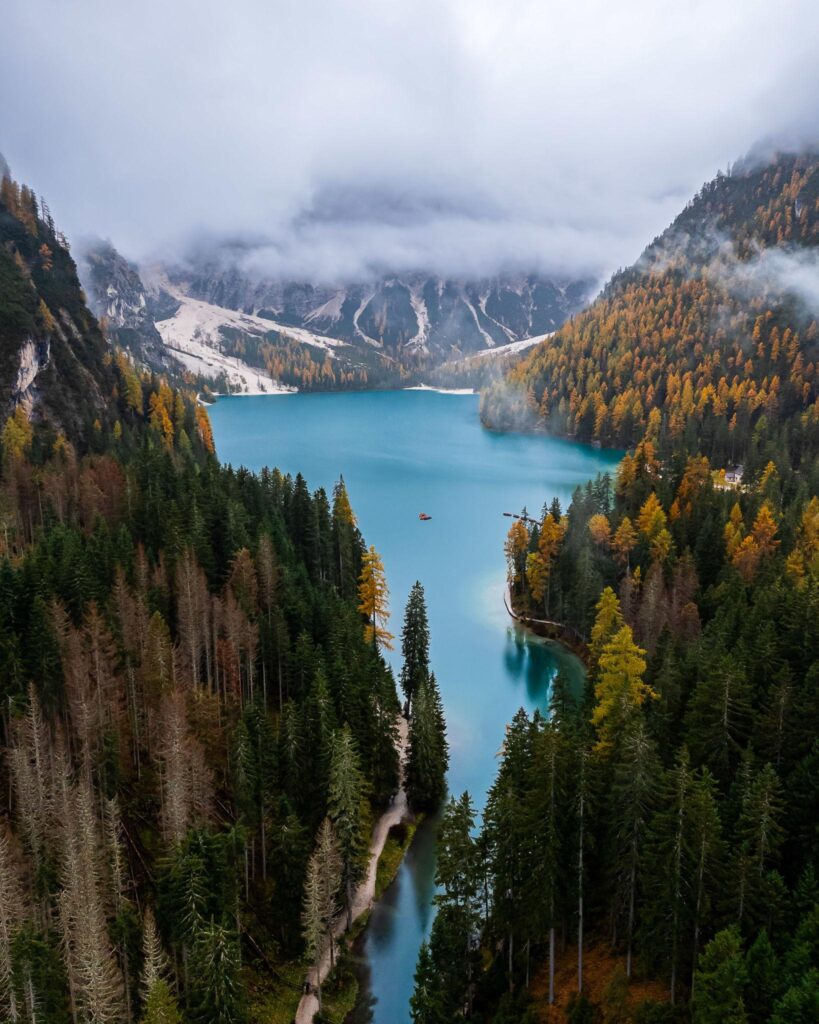Atmospheric aerial view of a turquoise alpine lake surrounded by forests and misty mountains in the Dolomites, Italy.