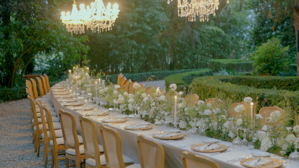 Why U.S. Couples Choose Italian Wedding Videographers 9 An outdoor luxury wedding reception featuring a long curved banquet table with white floral arrangements and crystal chandeliers hanging from trees in an Italian garden.