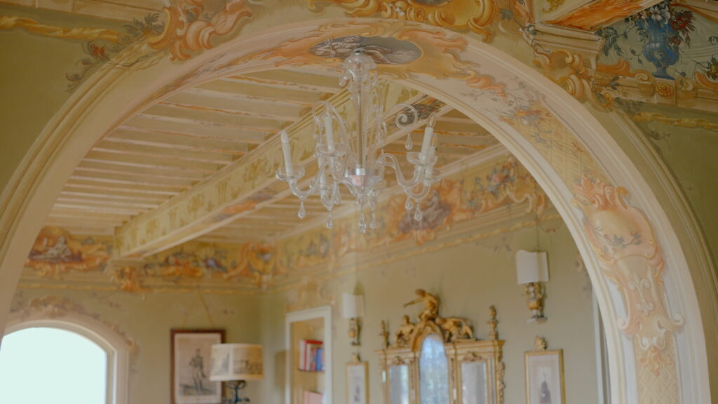 Why U.S. Couples Choose Italian Wedding Videographers 7 Interior view of a luxury Italian villa room showing detailed ceiling frescoes, a crystal chandelier, and antique gilded furniture through an open archway.
