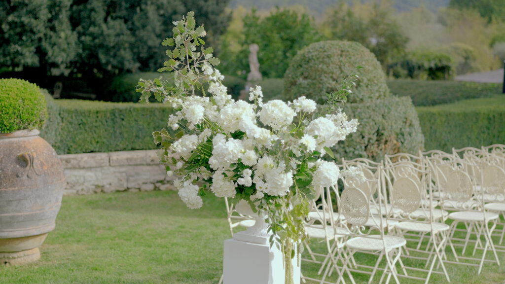 Why U.S. Couples Choose Italian Wedding Videographers 3 A horizontal image of an outdoor garden wedding setup. A large floral centerpiece in a white vase sits on a pedestal in front of empty rows of white wrought iron chairs. Formal gardens with hedges are in the background.