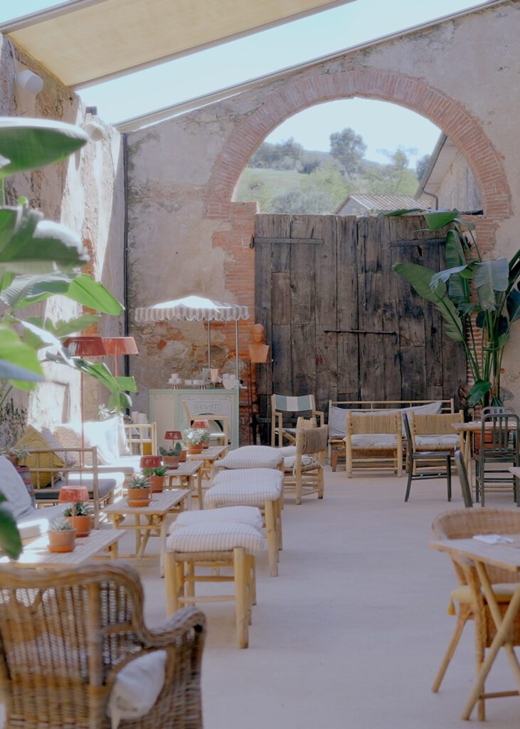 Rustic countryside lounge with wooden barn doors, woven furniture, tropical plants, and natural light inside a restored stone building.