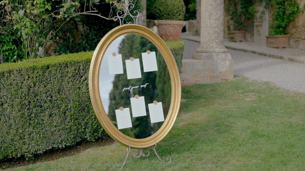 Why U.S. Couples Choose Italian Wedding Videographers 4 An ornate gold-framed oval mirror used as a wedding seating chart with wax-sealed cards, standing on a lawn in an Italian villa garden.